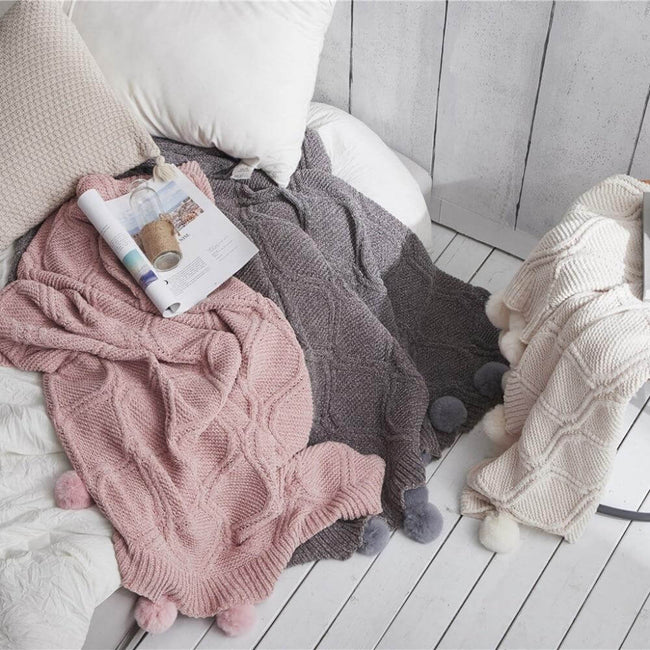 Three hygee knit pom-pom blankets laid out with pillows, a magazine, and a water bottle
