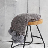 A gray hygee knit pom-pom blanket on a stool
