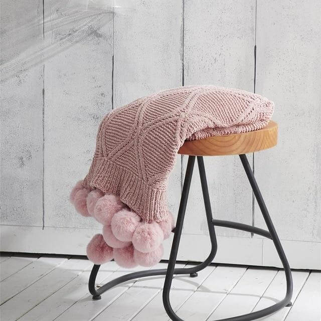 A pink hygee knit pom-pom blanket on a stool