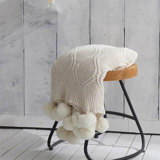 A white hygee knit pom-pom blanket on a stool