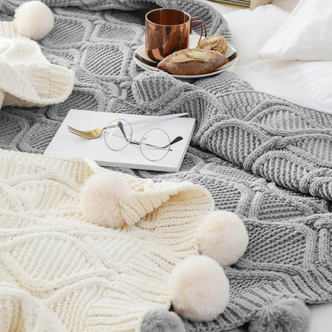 A white and a gray hygee knit pom-pom blanket on a bed with a mug