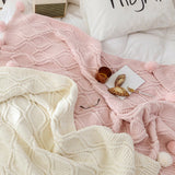 A white and a pink hygee knit pom-pom blanket on a bed with a mug