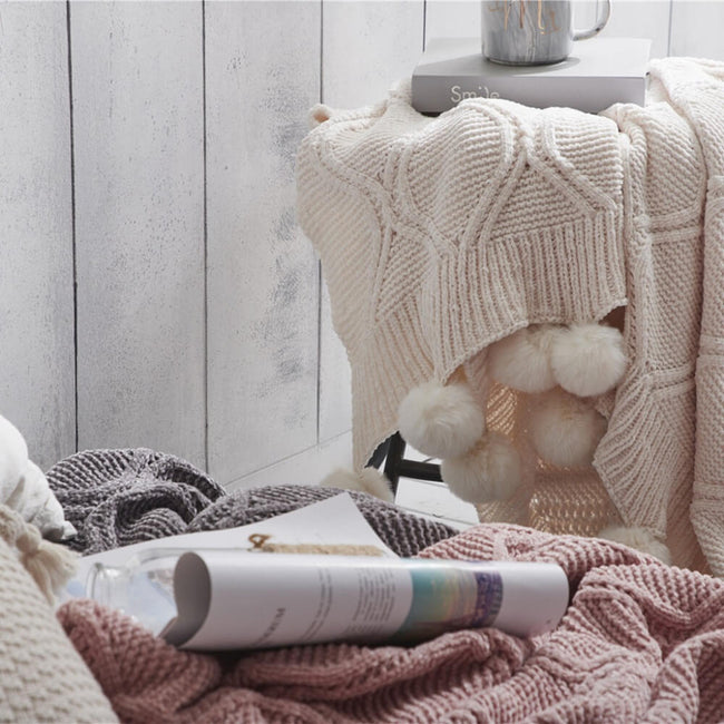Three hygee knit pom-pom blankets, white blanket on stool and other two on bed with magazine
